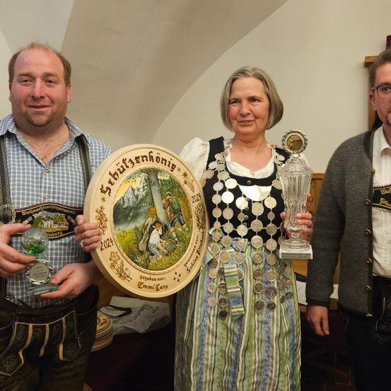 Drei Personen in traditioneller Kleidung halten Auszeichnungen; eine Frau hält eine große runde Auszeichnung und einen Glaspokal, ein Mann links von ihr hält einen Glaspokal und ein Mann rechts von ihr hält eine kleinere Glasauszeichnung.