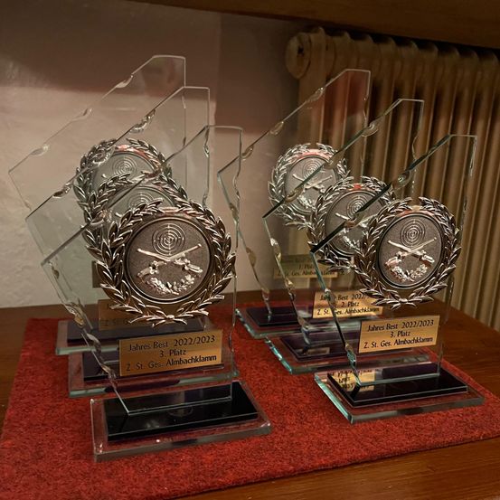 Vier Trophäen sind auf einem Tisch ausgestellt. Sie sind aus Glas mit silbernen Medaillen obenauf. Die Medaillen haben Inschriften wie 'Jahres Best 2022/2023' und 'Z. St. Ges. Almbachklamm'.