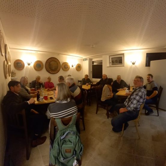 Eine Gruppe von Menschen sitzt um Tische in einem Restaurant, einige mit Brille. Der Raum hat einen Holzboden und Wände mit dekorativen Tellern.