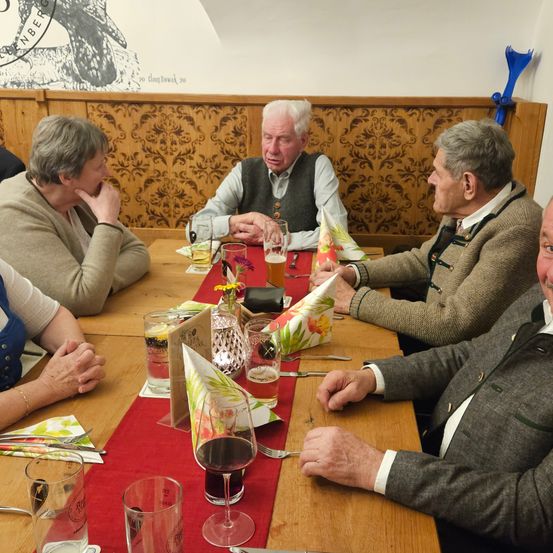 Eine Gruppe älterer Menschen sitzt in einem Restaurant um einen Tisch, trägt traditionelle Kleidung. Sie haben Getränke und Essen vor sich und einige haben Servietten. Die Wand hinter ihnen hat ein Design.