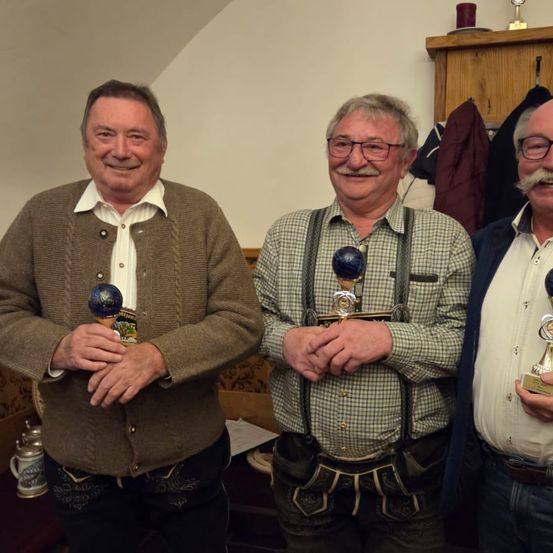 Drei Männer stehen lächelnd in einem Raum mit Holzregalen. Sie halten Trophäen. Der Mann links trägt einen Pullover, der in der Mitte ein kariertes Hemd mit Hosenträgern und der rechte einen Anzug.