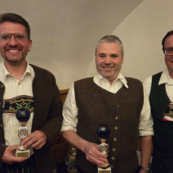 Drei Männer stehen lächelnd nebeneinander und halten Trophäen. Der Mann auf der linken Seite trägt eine Brille und eine Jacke mit 'Weihnachtsschutze'. Der Mann in der Mitte trägt eine braune Weste und hält eine Trophäe mit blauer Spitze. Der Mann auf der rechten Seite trägt eine grüne Weste und hält eine Trophäe.