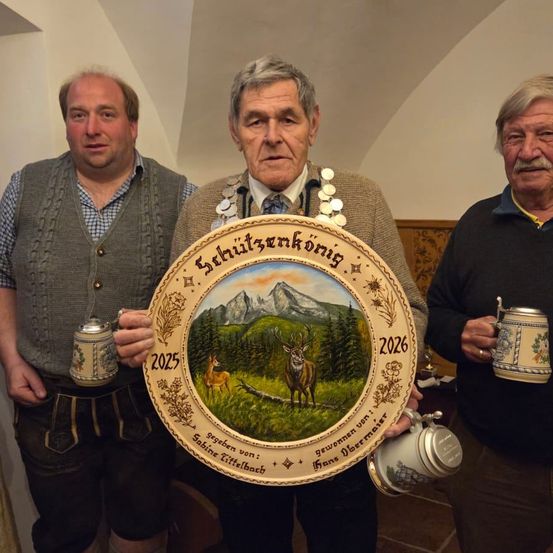 Drei Männer, einer hält einen dekorativen Teller mit einem Hirsch und Bergen, die anderen halten Tassen, stehen drinnen mit einer hölzernen Wand im Hintergrund.