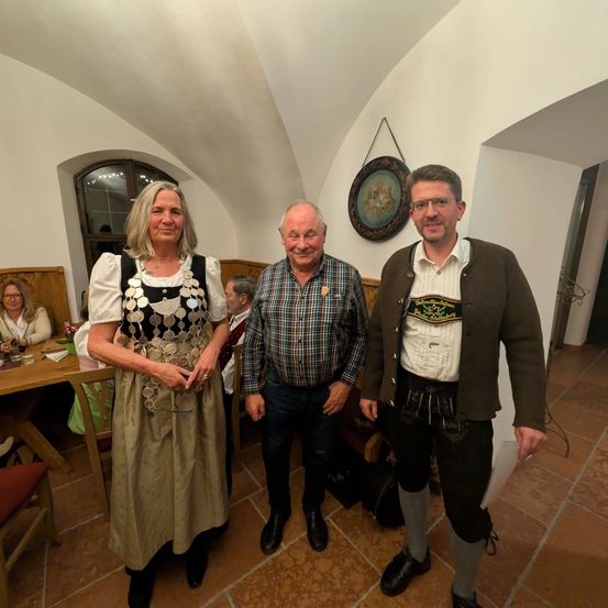 Drei Menschen stehen in einem Raum, zwei Männer und eine Frau, gekleidet in traditioneller Kleidung. Sie lächeln für die Kamera. Hinter ihnen steht ein Holztisch mit Blumen, ein Holzstuhl und ein Fenster mit Blick nach draußen.