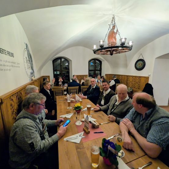 Eine Gruppe von Männern sitzt an einem Holztisch in einem Restaurant. Sie schauen auf ihre Telefone und halten Getränke. Der Tisch ist mit Gläsern, Blumen und Besteck gedeckt. Das Restaurant hat Holzwände, eine Kronleuchter und eine Wand mit Schrift.