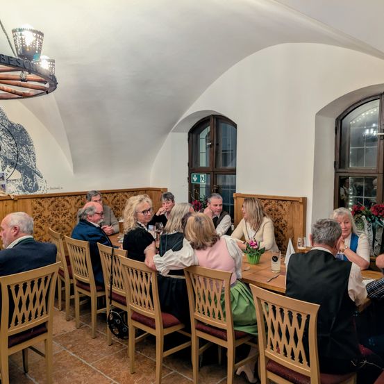 Eine Gruppe von Menschen sitzt um einen Tisch in einem Esszimmer. Der Tisch hat Blumen und Getränke. Der Raum hat Holzwände und Decken, eine Kronleuchter und ein Wandbild. Der Raum ist gut beleuchtet.