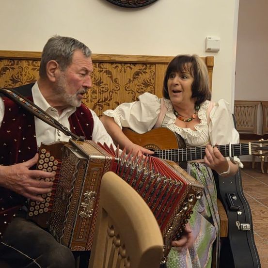 Ein älterer Mann und eine Frau sitzen lächelnd zusammen, während der Mann ein Akkordeon und die Frau eine Gitarre spielt. Hinter ihnen ist ein hölzerne Kopfstütze und ein Stuhl zu sehen.