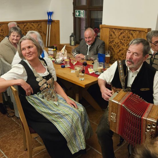 Mehrere Menschen sitzen in einem rustikalen Restaurant an einem Tisch und lächeln. Eine Frau in traditioneller Tracht sitzt gegenüber einem Mann, der ein Akkordeon spielt. Hinter ihnen lacht ein anderer Mann. Der Tisch ist mit Weingläsern, Tellern und einer Kerze gedeckt.