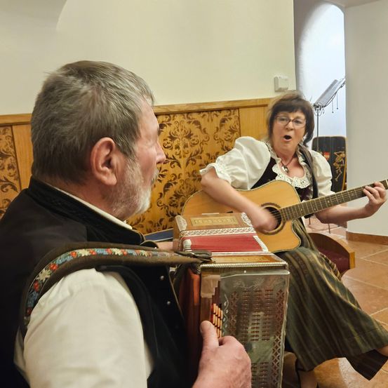 Ein Mann spielt Akkordeon, während eine Frau singt und Gitarre spielt. Sie sitzen auf einer Bank in einem Raum mit Holzwänden.