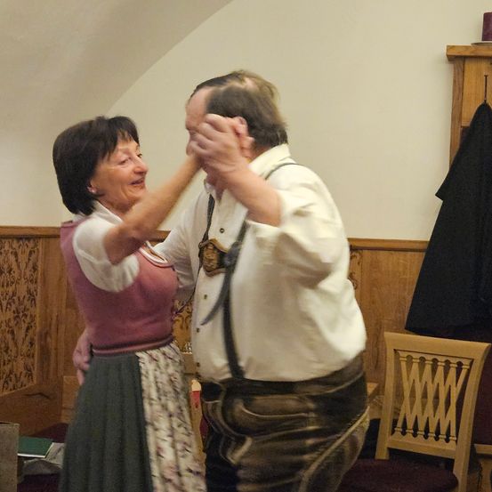 Eine ältere Frau zieht beim Tanzen an den Haaren eines Mannes, trägt ein rosa Oberteil und einen Blumenrock. Der Mann in traditioneller deutscher Kleidung scheint den Moment zu genießen.