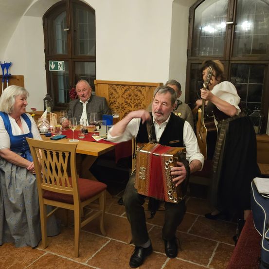 Eine Gruppe von Menschen sitzt in einem Restaurant. Ein älterer Mann spielt Akkordeon, während andere zusehen. Auf dem Tisch stehen Weingläser und eine Speisekarte.