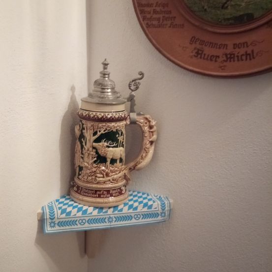 Ein Keramikkrug mit Hirschdesign steht auf einem blau-weißen Regal an einer weißen Wand. Daruber befindet sich ein runder brauner Teller mit fremdsprachigem Text.