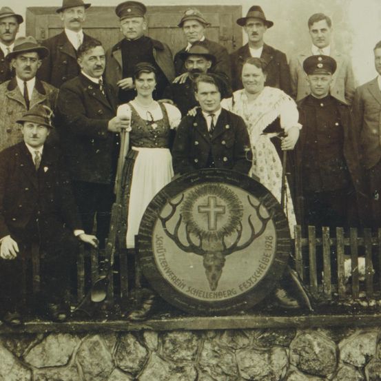 Ein altes Foto zeigt eine Gruppe von Menschen in vintage Kleidung, einige in Uniformen, die hinter einem Holzzaun posieren. Ein rundes Emblem mit einem Hirschschädel und einem Kreuz ist in der Mitte.