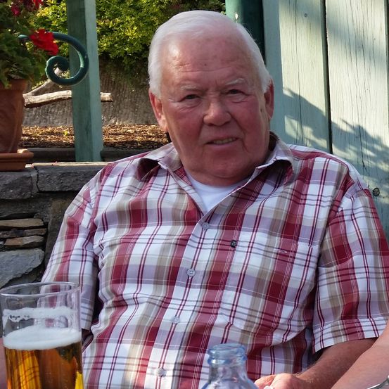 Ein älterer Mann mit weißen Haaren sitzt im Freien und trägt ein rot-weiß kariertes Hemd. Vor ihm steht ein Glas Bier. Dahinter befindet sich ein Holzzaun und eine Topfpflanze.