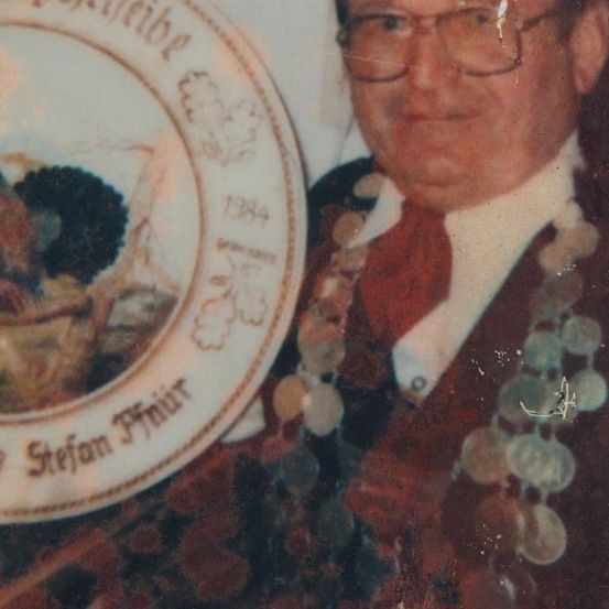 Ein älterer Mann mit Brille und Halskette hält einen Teller mit einer Zeichnung eines Korbes und Trauben und dem Namen 'Stefan Fmuir' darauf.