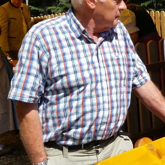 Ein älterer Mann in einem blau-rot-weiß karierten Hemd steht neben einem Holzzaun.