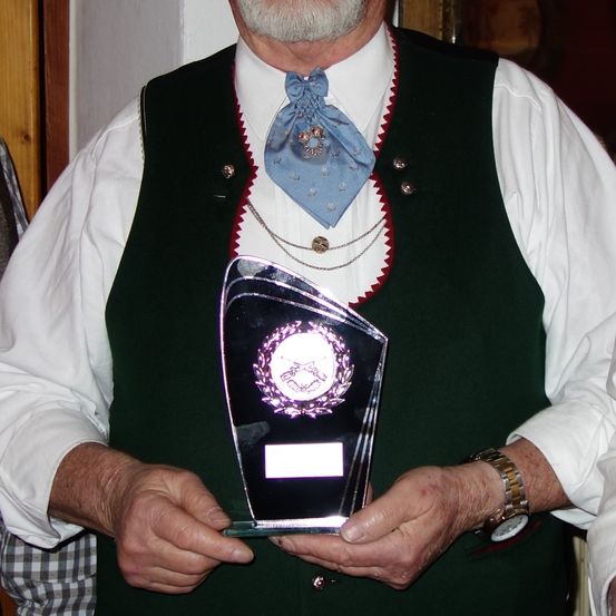 Ein älterer Mann in traditioneller Kleidung hält eine Trophäe mit einem Gewehremblem und weißer Schrift. Er trägt eine blaue Fliege und hat eine Uhr am Handgelenk.