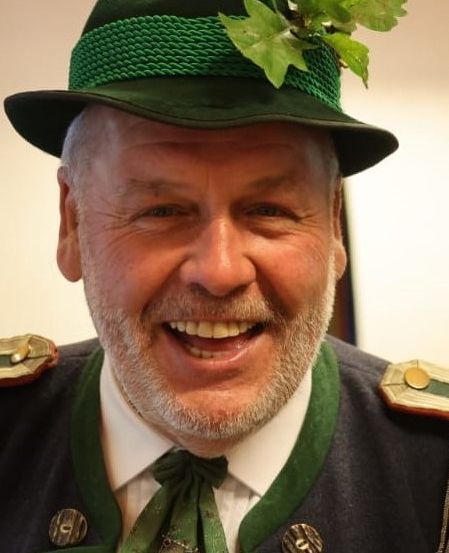 Ein Mann mit einem grünen Hut, der mit Blättern verziert ist, einem grünen und goldenen Kostüm und einer grünen Krawatte mit einer dekorativen Brosche. Er lächelt.