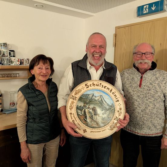 Drei Menschen stehen zusammen in einem Raum, lächeln und posieren für ein Foto. Der Mann in der Mitte hält einen runden Teller mit einer malerischen Landschaft. Die Frau auf der linken Seite trägt eine Weste und der Mann auf der rechten Seite trägt eine Brille. Hinter ihnen ist eine Holztür mit einem Regal mit Tassen und Gläsern.
