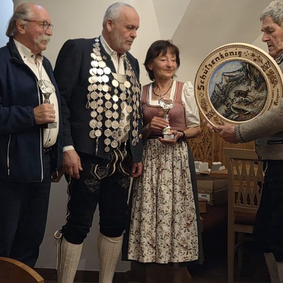 Vier Personen, gekleidet in traditioneller Kleidung, stehen zusammen. Ein Mann auf der linken Seite hält eine Trophäe. Eine Frau in der Mitte hält eine Trophäe und einen dekorativen Teller. Ein Mann auf der rechten Seite hält einen dekorativen Teller. Hinter ihnen steht ein Holzstuhl und ein Tisch.
