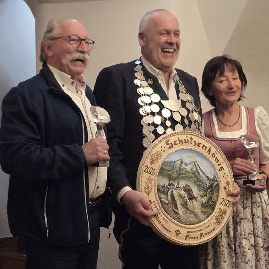 Drei Personen stehen eng beieinander, lächeln und halten Pokale. Der Mann in der Mitte trägt eine Kette von Medaillen, und ein gerahmtes Kunstwerk ziert die Mitte. Die Frau rechts trägt ein traditionelles Kleid.