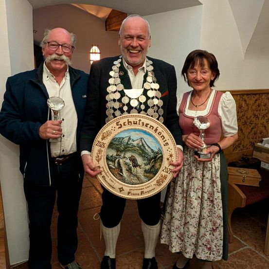 Drei Menschen stehen lächelnd zusammen. Ein Mann auf der linken Seite hält einen Pokal, ein anderer Mann in der Mitte hält eine große dekorative Platte und eine Frau auf der rechten Seite hält ebenfalls einen Pokal. Sie sind alle in traditioneller Kleidung gekleidet.