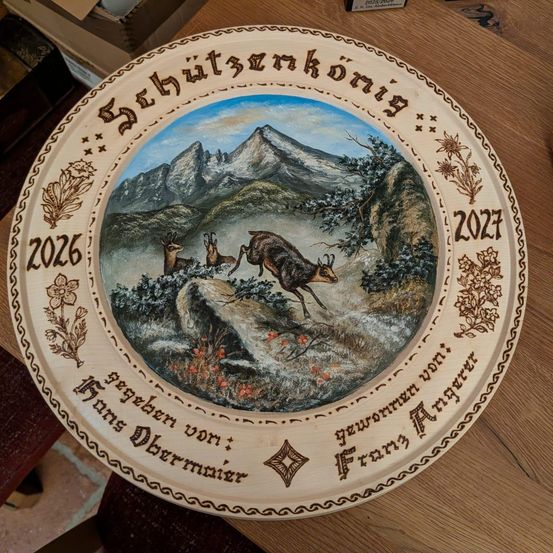 Eine Holzplatte mit den Worten 'Schutzkonig' und '2026'. Im Zentrum befindet sich ein Bild von flüchtenden Hirschen in den Bergen. Die Platte ist von Franz Amsler signiert.