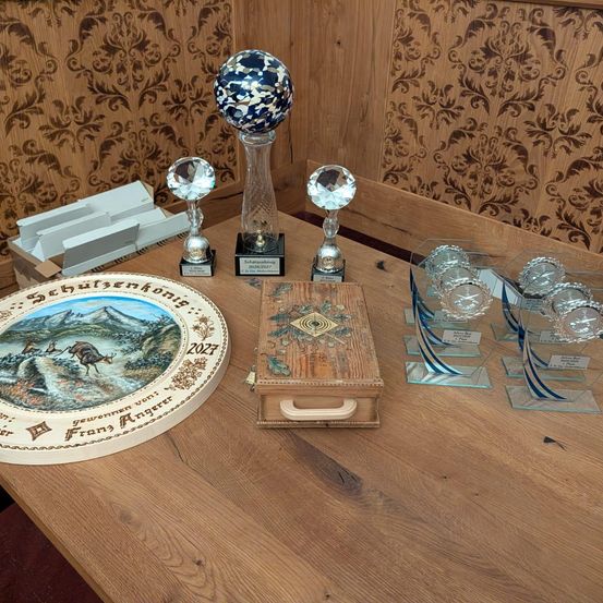 Ein Holztisch zeigt Trophäen, eine Schachtel und einen dekorativen Teller mit Bergen. Die Trophäen sind aus Kristall und Glas. Die Schachtel hat ein geometrisches Design. Der Teller zeigt einen Hirsch und eine Berglandschaft.