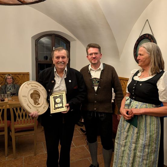 Drei Menschen stehen lächelnd zusammen in einem Raum mit einem hölzernen Fenster. Der Mann auf der linken Seite hält ein gerahmtes Foto und eine kleine Plakette. Der Mann in der Mitte trägt eine Brille und einen braunen Pullover. Die Frau auf der rechten Seite trägt ein traditionelles Kleid. Hinter ihnen steht ein Tisch mit Stühlen.