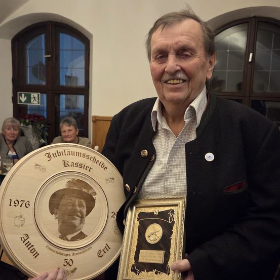Ein älterer Mann hält eine Plakette und einen gerahmten Preis, lächelt. Die Plakette zeigt ein Porträt eines Mannes aus dem Jahr 1976 mit einer 50-jährigen Jubiläumsinschrift.