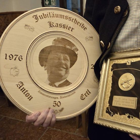 Eine Person hält einen Holzteller und eine gerahmte Auszeichnung. Der Teller trägt die Aufschrift 'Jubilaumsscheibe Kassierer Anton' mit dem Jahr 1976 und '50'. Die Auszeichnung hat ein goldenes Design und Text.