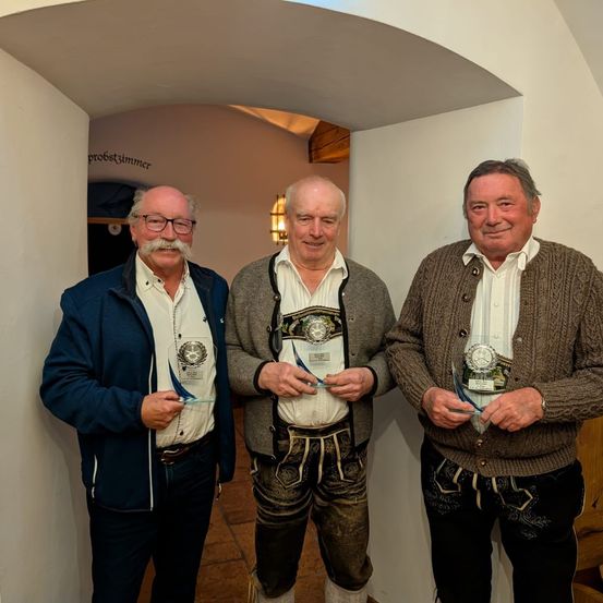 Drei Männer stehen in einer Türöffnung und lächeln. Jeder hält eine Plakette. Der linke Mann trägt eine Jacke und eine Brille. Der mittlere Mann trägt einen Pullover und traditionelle Kleidung. Der rechte Mann trägt einen Pullover und traditionelle Kleidung. Hinter ihnen ist eine Wand mit Text.