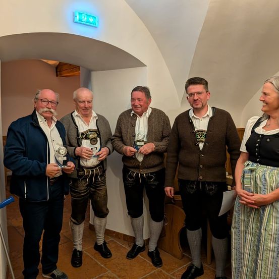 Fünf Personen, vier Männer und eine Frau, stehen in einem Raum und lächeln für die Kamera. Sie tragen traditionelle Kleidung. Der Mann auf der linken Seite trägt eine blaue Jacke, während die anderen braune Pullover und Hosen tragen. Sie halten kleine Trophäen.