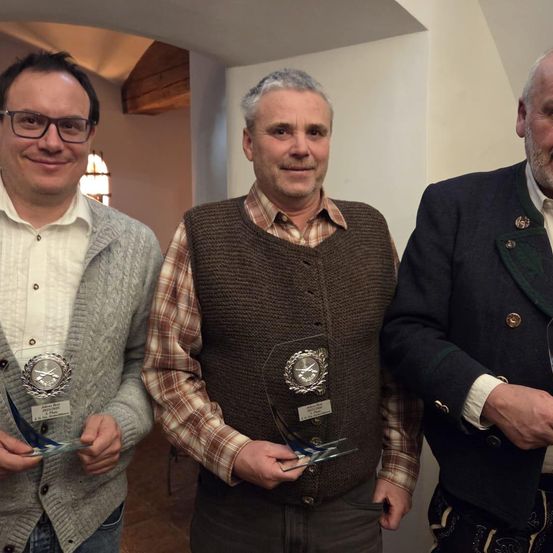 Drei Männer stehen nebeneinander und halten Auszeichnungen. Der Mann auf der linken Seite trägt einen grauen Pullover und eine Brille. Der Mann in der Mitte trägt eine braune Weste und ein kariertes Hemd. Der Mann auf der rechten Seite trägt einen schwarzen Mantel mit grünem Kragen.