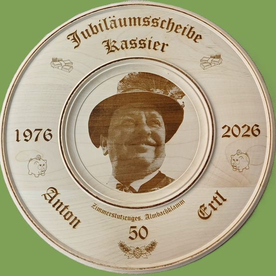 Eine hölzerne Gedenktafel zeigt ein Porträt eines Mannes mit Hut. Sie markiert seinen 50. Jahrestag von 1976 bis 2026. Ein Sparschwein und Tannenzapfen schmücken die Tafel.