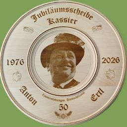 Eine hölzerne Gedenktafel zeigt ein Porträt eines Mannes mit Hut. Sie markiert seinen 50. Jahrestag von 1976 bis 2026. Ein Sparschwein und Tannenzapfen schmücken die Tafel.