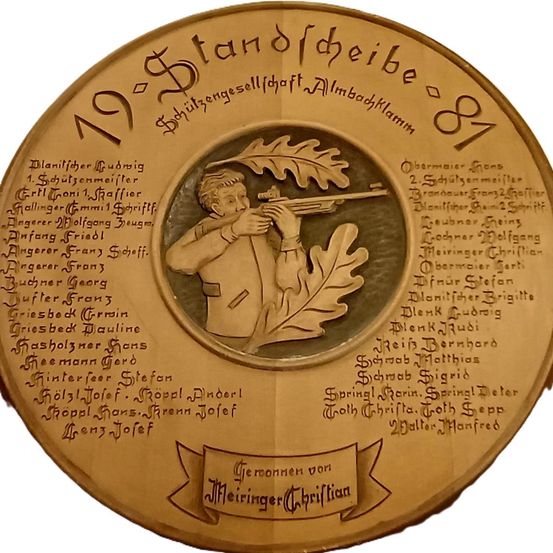 Eine kreisförmige Plakette zeigt einen Mann mit einem Gewehr und einem Eichblatt, umgeben von Namen und Titeln in deutscher Sprache. Die Plakette ist goldfarben und hat ein historisches, formelles Aussehen.