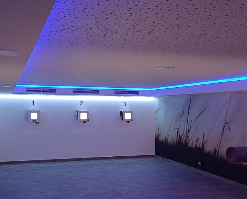 Ein leerer Raum mit weißen Wänden und einer lila Decke. Drei nummerierte Paneele sind an der Wand montiert. Ein landschaftliches Wandbild schmückt die rechte Wand.