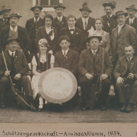 Ein Schwarz-Weiß-Foto einer Gruppe von Menschen, hauptsächlich Männern, die Hüte und Krawatten tragen. Das Bild hat ein Wasserzeichen, das 'Schützengesellschaft - Almbachklamm, 1934' lautet.