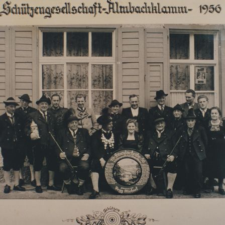 Eine Gruppe von Menschen in traditioneller Kleidung steht vor einem Gebäude. Die Gruppe posiert für ein Foto mit einem Schild in der Mitte. Das Foto hat ein Wasserzeichen, das Schutzengesellschaft Almbachklamm 1956 lautet.