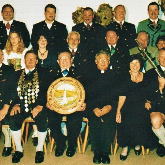 Eine Gruppe von Menschen in traditioneller Kleidung posiert für ein Foto, einige tragen Medaillen und halten eine goldene Plakette mit einem Logo.