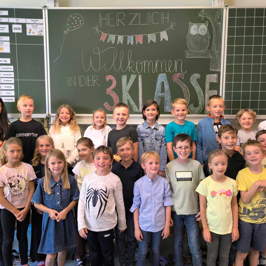 Eine Gruppe von Schülern in einem Klassenzimmer lächelt für ein Foto vor einer Tafel, auf der Herzlich Willkommen in der 3 Klasse steht.