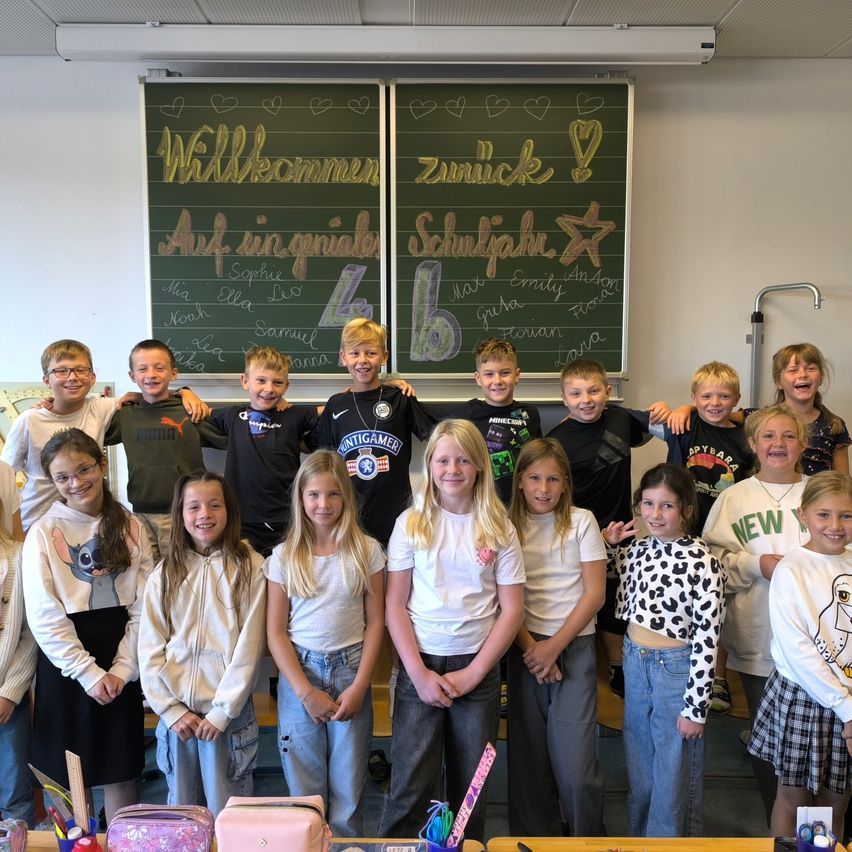 Eine Gruppe von Kindern posiert vor einer Tafel mit deutscher Beschriftung. Sie lächeln und tragen verschiedene Kleidungsstücke.