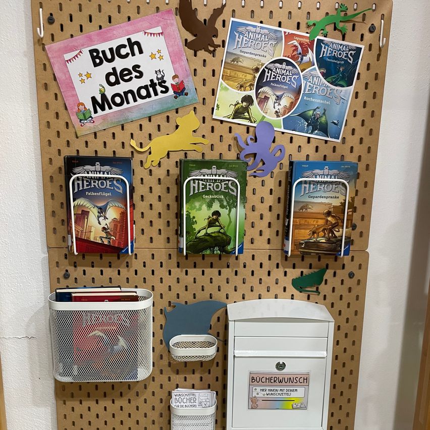 Ein Anschlagbrett mit verschiedenen Büchern und Materialien. Es gibt einen Bereich für das 'Buch des Monats', mit Büchern in bunten Umschlägen und Illustrationen. Zusätzliche Bücher sind in einem hängenden Fach ordentlich organisiert. Es gibt auch einen weißen Kasten mit der Aufschrift 'Wunschbuch'.