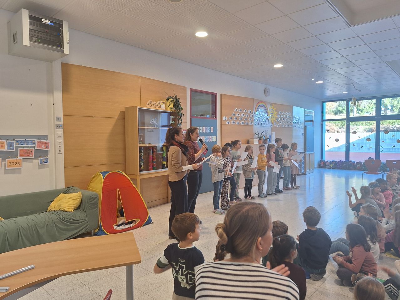 Eine Gruppe von Kindern und zwei Frauen steht in einem Klassenzimmer, hält Papiere und liest. Eine Frau hält ein Mikrofon, und ein Zelt ist im Hintergrund.