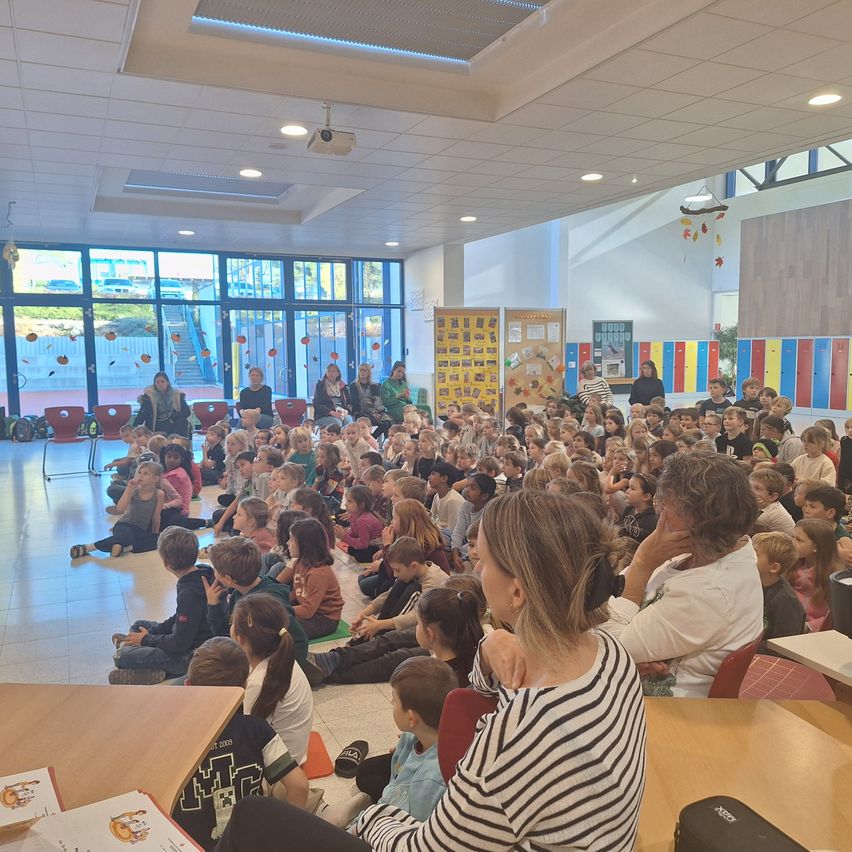 In einem großen Klassenzimmer sitzen eine Gruppe von Kindern auf dem Boden und lauschen einer Frau. Hinter ihnen sitzen weitere Kinder auf Stühlen. Der Raum hat große Fenster mit farbenfrohen Dekorationen und eine Projektorlampe an der Decke.