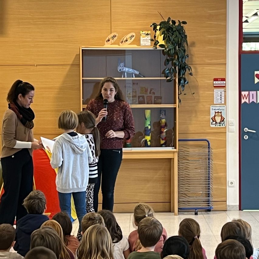 Zwei Frauen sprechen eine Klasse voller Kinder an. Eine hält ein Mikrofon, die andere ein Buch. Das Klassenzimmer hat Holzwände, ein Bücherregal und eine Tür mit Herz- und Eulendekoration.