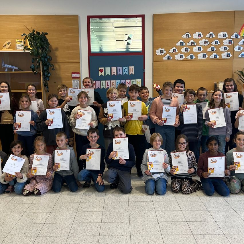 Eine Gruppe von Kindern und Erwachsenen versammelt sich in einem Klassenzimmer und hält Urkunden. Sie lächeln und posieren für ein Foto mit einer Lehrerin.
