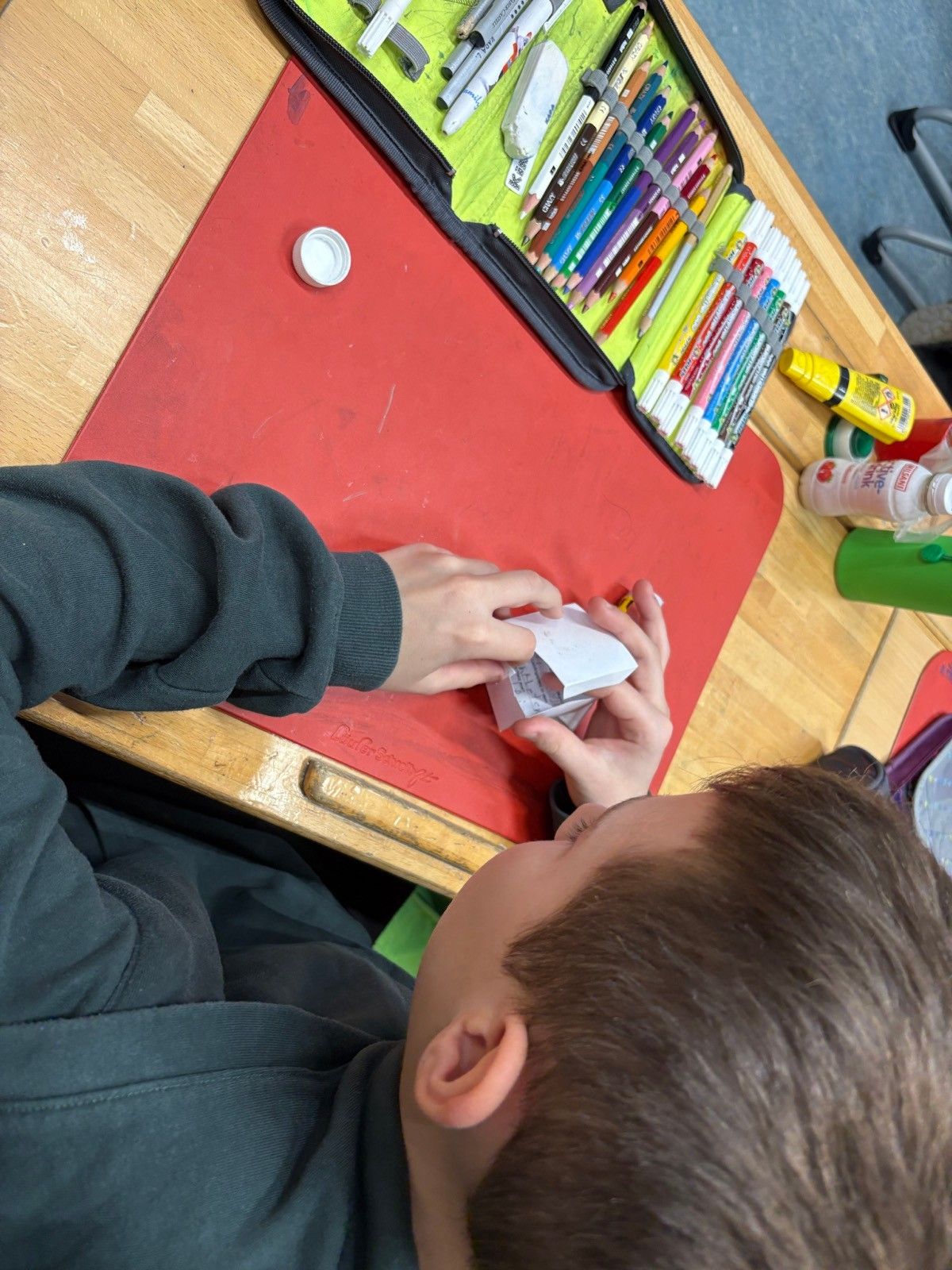 Ein Junge arbeitet mit einem Bleistift an einer Papierbastelei, sitzt an einem Holzschreibtisch mit einer roten Unterlage und verschiedenen Bastelutensilien um ihn herum.