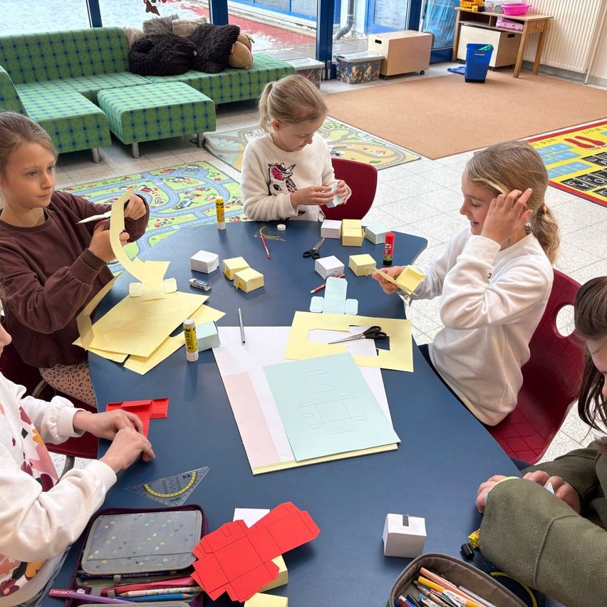 Fünf junge Mädchen sitzen um einen Tisch in einem Klassenzimmer und beschäftigen sich mit einer Bastelaktivität. Sie haben verschiedene farbige Papiere und Materialien auf dem Tisch verteilt.
